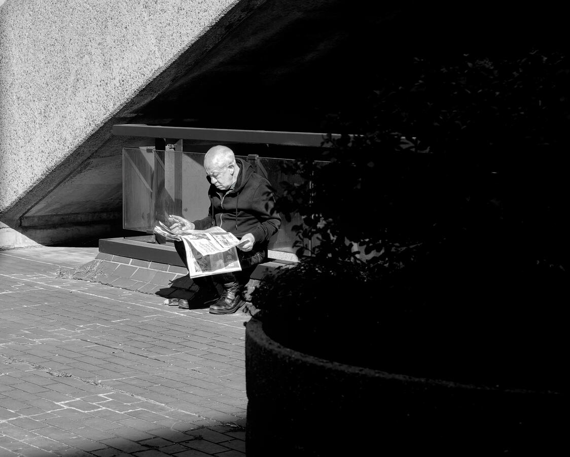 A man sitting on a kerbside reading a newspaper whilst smoking a cigarette