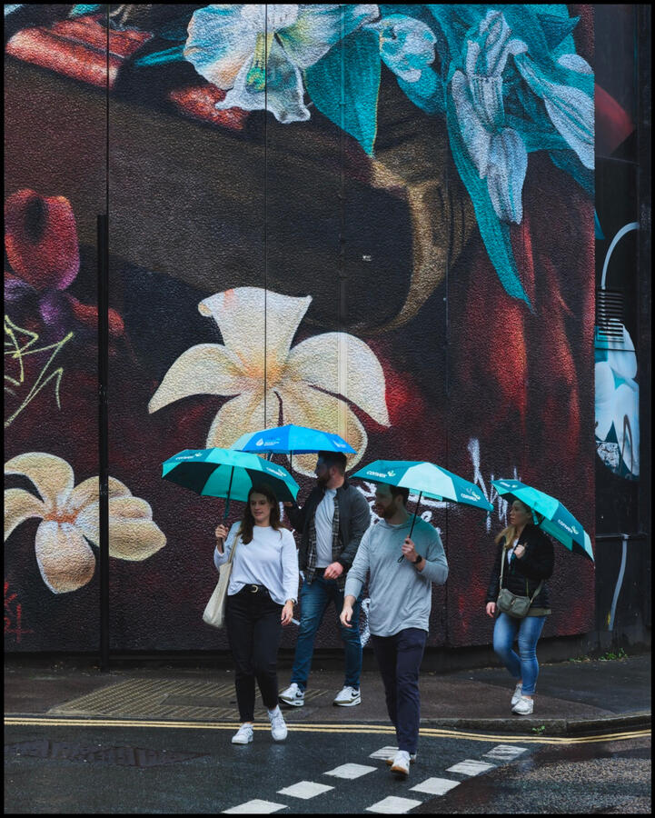 People crossing road with bright blue umbrellas with large blue mural on wall behind them